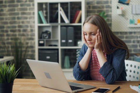 A young woman sitting at a desk looking frustrated while staring at a laptop screen, resting her head on her hands. 